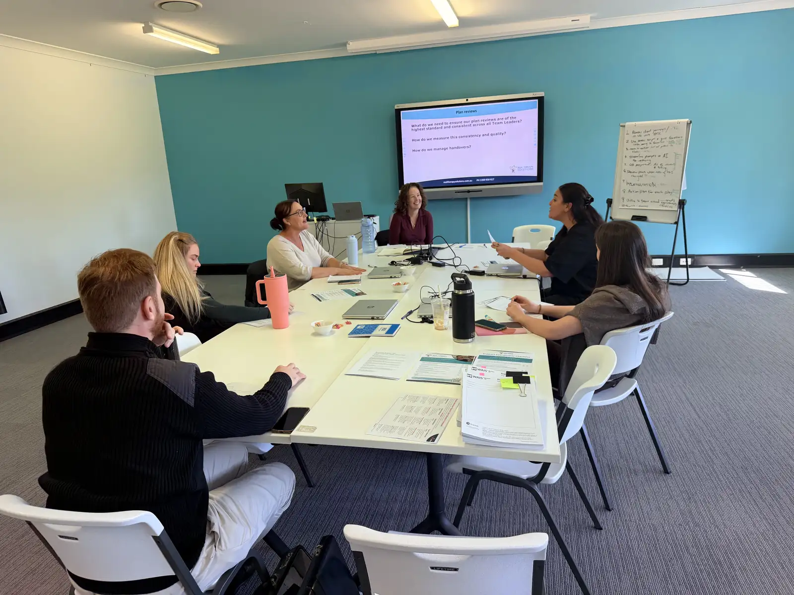 Real Therapy Solutions team seated around a boardroom table discussing plan reviews, with a presentation slide and whiteboard notes on the wall