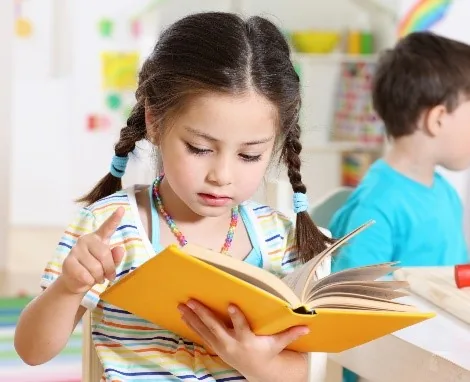 A girl reading an open book at a desk beside a classmate