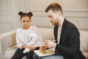 A child holding a plush toy listens to a man in a therapy session