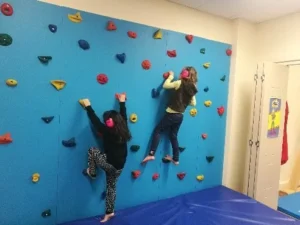 Two children climbing a blue indoor climbing wall above a crash mat