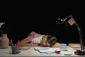 Tired woman asleep on a desk with office supplies under a lamp in dark workspace