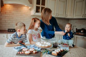 Children and woman in casual clothes at table with tasty desserts with cream on trays near jars in light kitchen