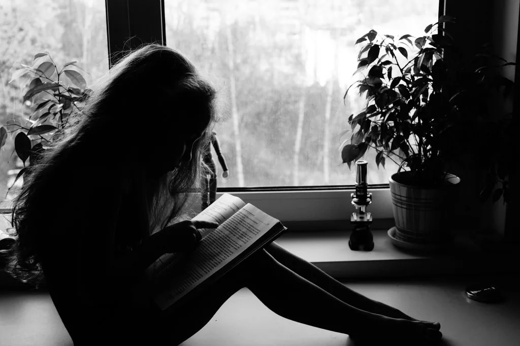 Black and white photograph of a person reading a book beside a window